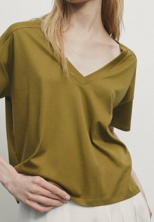Kobieta w oliwkowo zielonym T-shircie z krótkim rękawem i dekoltem w serek, lekko wpuszczonym w białą plisowaną spódnicę, ręka oparta na biodrze.