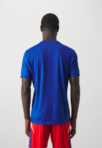 Maglietta atletica a maniche corte da uomo in un blu vivace, caratterizzata da una texture liscia e un collo a giro. Indossata con pantaloni corti rossi, mostra uno stile sportivo casual.