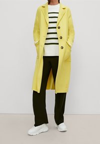 Manteau long jaune avec de gros boutons et des poches, superposé sur un pull rayé blanc et vert, associé à un pantalon noir et des baskets blanches.