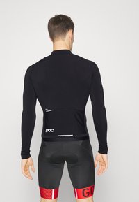 Maillot de cyclisme à manches longues en noir avec un design ajusté, comprenant une poche arrière, un logo, une texture lisse et des accents rouges contrastants sur le short.