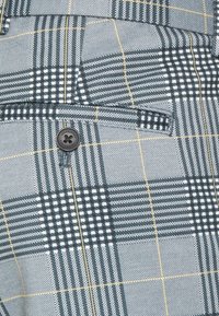 Šedá kostkovaná látka s tenkými černými a žlutými pruhy, obsahující kapsu na klopu s knoflíkem, na kterém je vyraženo "Jack & Jones".