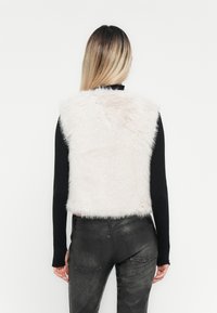 Gilet en fausse fourrure rose pâle avec une texture courte et douce, associé à un haut noir à manches longues et un pantalon noir ajusté.