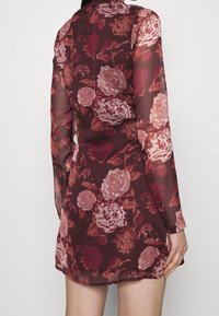 Blommig klänning i genomskinlig burgundy-tyg med långa ärmar, med stora mönster av rosa och krämfärgade rosor. Inkluderar en subtil detalj med öppning i ryggen.