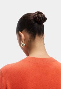 Pomarańczowy sweter dzianinowy z okrągłym dekoltem, widziany z tyłu. Włosy ułożone w warkocz w koku, z dużymi złotymi kolczykami.