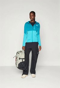 Naine kannab sinist lukuga kapuutsi ja musti kottpüksi, hoides halli ja musta seljakoti, seistes ühtlasel valgel taustal.