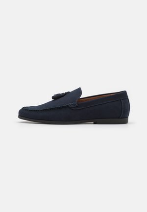 Mocasín masculino de ante azul marino con detalle de borlas, suela plana negra y costuras visibles a lo largo de los bordes, mostrado de perfil.