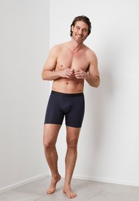 Dunkelblaue Boxer-Briefs mit glatter Textur und elastischem Taillenbund, getragen von einem männlichen Modell, das barfuß vor einer hellen Wand steht.