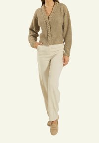 Beige gestrickte Cardigan mit tiefem V-Ausschnitt, strukturiertem Muster und geflochtenen Details. Kombiniert mit hellbeigen Leinenhosen und Slippern.