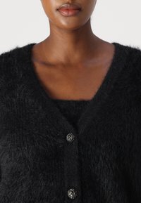 Czarny puszysty kardigan z głębokim dekoltem w kształcie V, ozdobiony trzema dekoracyjnymi guzikami wysadzanymi cyrkoniami, podkreślającymi jego miękką teksturę.