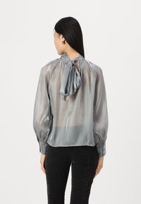 Blusa di raso argento con colletto alto e dettaglio a fiocco, maniche a sbuffo lunghe e aspetto trasparente. Indossata con pantaloni neri testurizzati.