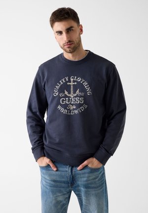 Joven con una sudadera azul marino con un ancla y el texto "Ropa de Calidad Guess Estilo Mundial", combinado con vaqueros azules, con las manos en los bolsillos.