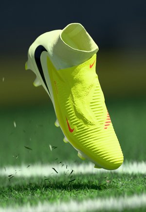 Scarpette da calcio di un vivace giallo con superficie testurizzata, dettagli bianchi e neri, e design high-top. Visibile detrito da campo sotto la scarpina.