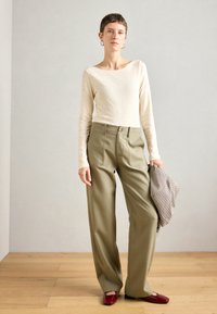 Grietos ilgomis rankovėmis marškiniai, alyvuogių spalvos plačios kelnės ir raudoni plokšti batai. Laiko raštuotą, tekstūrinę švarką. Natūralaus medžio grindų fono.