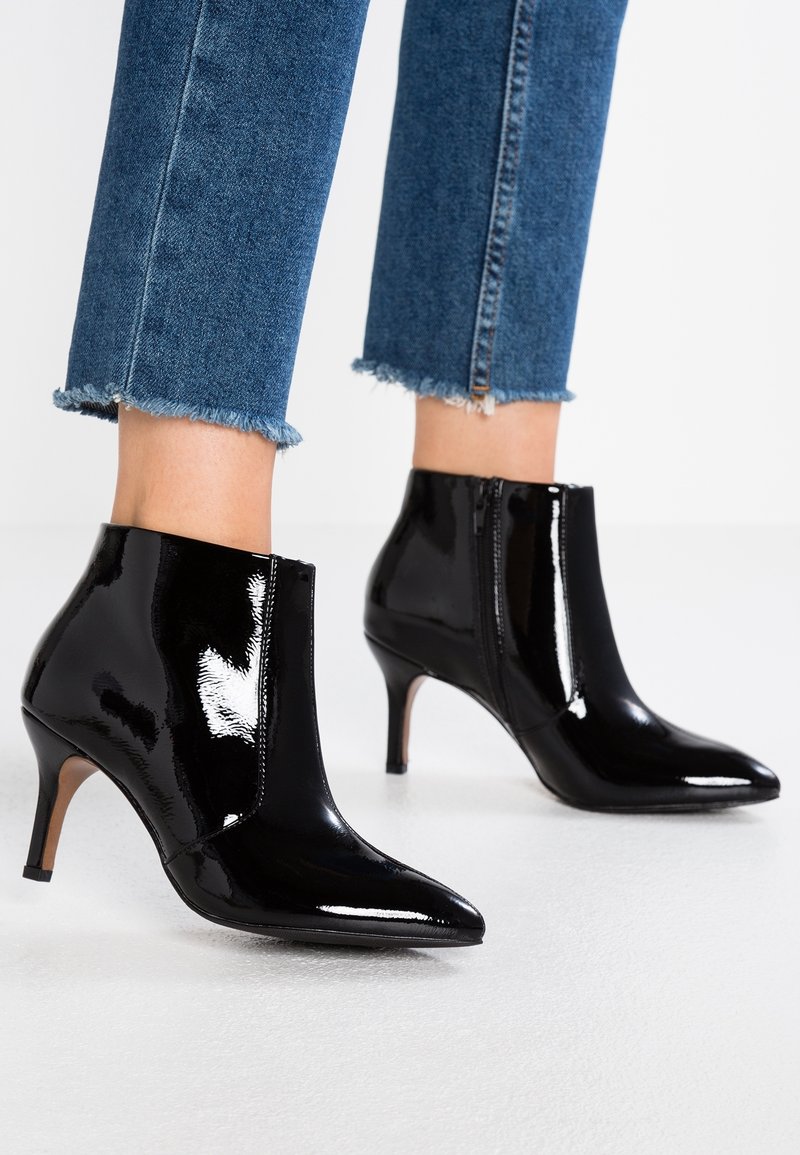 Personne portant des bottines en cuir verni noir brillant, à bouts pointus et talons fins, associées à un jean bleu à ourlet effiloché.