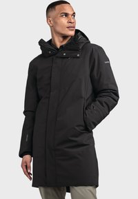 Schwarzer langer Mantel mit einem hohen Kragen und verstellbarem Kapuze. Aus wasserabweisendem Material gefertigt, mit seitlichen Taschen und modernem, schlankem Design.