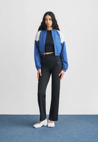 Veste courte bleue et blanche portée sur un haut noir, associée à un pantalon évasé noir et des baskets blanches. La tenue arbore la marque Nike.