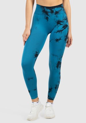 Femme portant des leggings bleus taille haute avec des motifs noir tie-dye et des baskets blanches, se tenant devant un fond gris uni.
