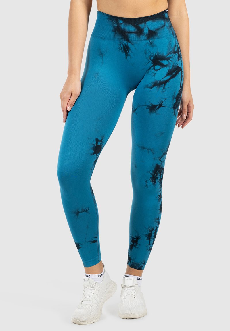 Kobieta w niebieskich leginsach z wysokim stanem w czarne wzory tie-dye i białych trampkach, stojąca na jednolitym szarym tle.