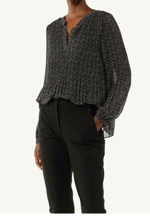 Person wearing black patterned long-sleeve blouse with smocked chest and black pants, hand in pocket, plain light background.