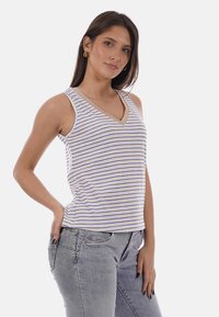 Femme portant un haut blanc sans manches avec de fines rayures bleues et des bordures beiges, associée à un jean gris clair, debout avec une main dans la poche.