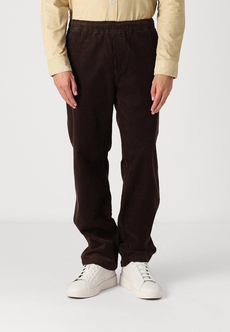 Homme portant un pantalon en velours côtelé marron foncé, une chemise beige et des baskets blanches, debout devant un fond clair uni.