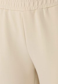 Beige stretchig tyg som visar en närbild av byxmaterial med ett elastiskt midjeband och en slät textur, med subtila veck.