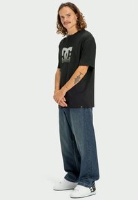 Tricou negru cu mânecă scurtă, având un logo mare gri DC pe față, asociat cu blugi lejeri din denim albastru și adidași albi.