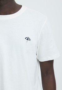 Witte katoenen t-shirt met een klein marineblauw geborduurd paraplu-motief op de linkerborst. Korte mouwen en ronde hals.