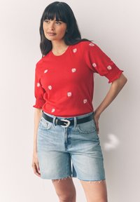 Young woman wearing red knit top with white daisy prints, light denim cutoff shorts, and black belt, standing against plain white background.