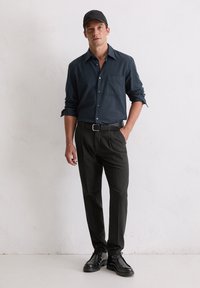 Overhemd met knopen in donkerblauw met een zak, zwarte broek, zwarte schoenen en een zwarte pet. Soepele stof en getailleerde pasvorm.