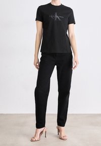 Czarna bawełniana koszulka z krótkim rękawem i srebrnym logo, połączona z czarnymi prostymi dżinsami i nagimi sandałami na obcasie. Gładka tekstura.