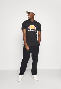 Camiseta negra con un logo gráfico colorido, combinada con pantalones negros y zapatillas blancas, además de una gorra blanca llevada de lado.