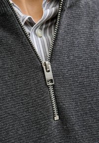 Hallhalli, hallhalli tõmblukuga kampsun tekstuurmaterjalist, millel on hõbedane tõmblukk. Allpool on osaliselt nähtav valge särk, millel on õhukesed hallid triibud.