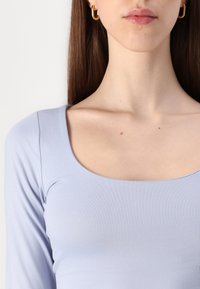 Lyseblå langermet topp med firkantet utringning og jevn, elastisk tekstur. Har en tettsittende design som fremhever kragebeinet.