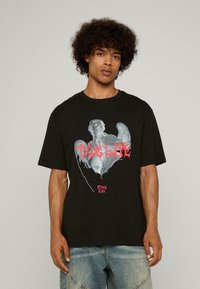 Čierne bavlnené tričko s grafický potlačou postavy a textom "Thug Life" v červenej farbe, krátke rukávy a voľný strih.