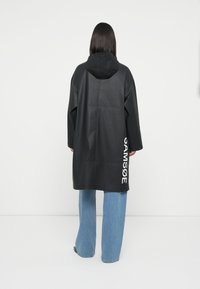 Black waterproof coat with a hood, featuring a loose fit and white brand lettering on the side. Worn over light blue wide-leg jeans.