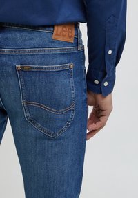 Calças de ganga azul com um emblema de couro castanho da marca Lee na cintura. As características incluem cinco bolsos e detalhes de costura contrastantes.