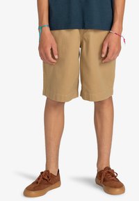 Shorts kaki à coupe droite, avec ourlet retroussé et tissu robuste. Portés avec des baskets en daim marron et agrémentés de bracelets colorés.