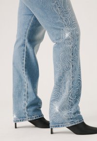 Jean en denim bleu clair avec une coupe décontractée, ornés de motifs tourbillonnants en strass sur les côtés et terminés par une légère frange.