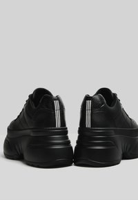 Svarta sneakers med strukturerade läderöverdrag, tre vita ränder på baksidan och en kraftig sula med ett räfflat mönster.