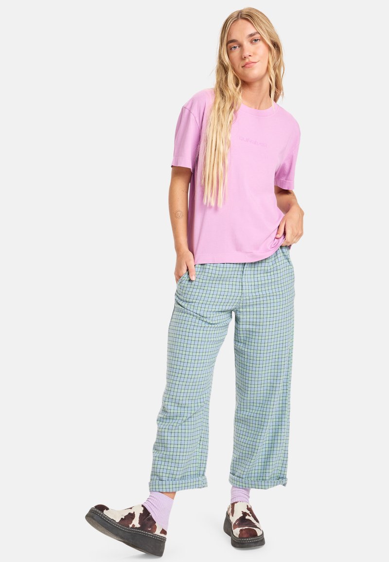 Roze katoenen t-shirt, ontspannen pasvorm. Lichtblauwe geruite broek met een losse snit. Bruine schoenen met een koeienprint en roze sokken.