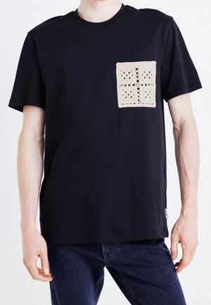 Person trägt ein schwarzes T-Shirt mit kurzen Ärmeln, auf der linken Brust ein beiges, gehäkeltes Quadratmuster und dunkelblaue Jeans.