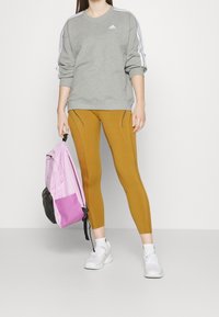 Kvinna som håller en rosa och lila ryggsäck, klädd i en grå Adidas-tröja, senapsgula leggings, vita strumpor och vita sneakers mot en enfärgad vägg.