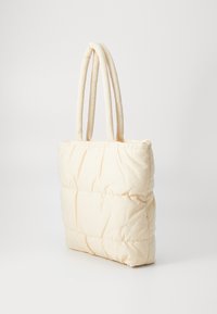 Sac fourre-tout matelassé en tissu beige clair et doux. Dispose de deux poignées rembourrées, d'une forme rectangulaire et d'un détail de couture visible sur la surface.