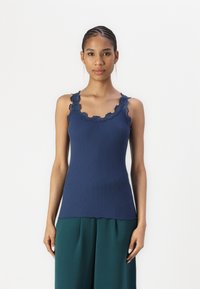 Marineblauw geribd tanktop met golfrand kant afwerking langs de halslijn en schouderbanden. Het aansluitende ontwerp contrasteert met de losse groene broek.