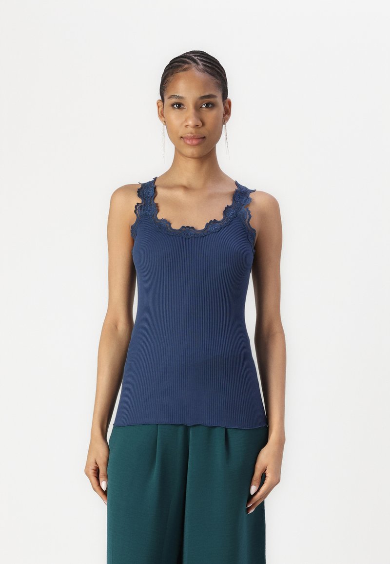 Marineblauw geribd tanktop met golfrand kant afwerking langs de halslijn en schouderbanden. Het aansluitende ontwerp contrasteert met de losse groene broek.