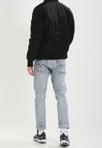 Veste bomber noire à texture lisse, poignets côtelés et devant zippé. Jeans bleu clair à coupe classique avec revers roulés. Baskets aux couleurs sombre et claire.