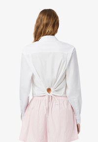 Camicia bianca con schiena arricciata fissata da un foro circolare e cordoncino. Abbinata a una gonna striata bianca e rosa chiaro.