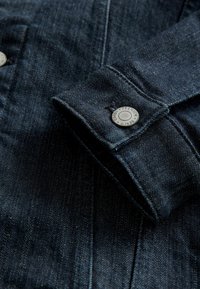 Dark blue denim jacket sleeve with visible silver button labeled "Premium Denim Goods" on cuff.