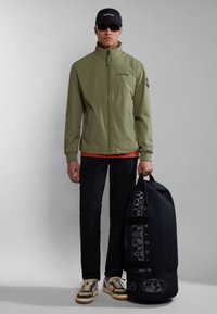 Giacca a zip verde oliva, cappellino nero e pantaloni scuri; tiene una grande borsa blu navy con logo. Le scarpe sono beige con accenti verdi e arancioni.